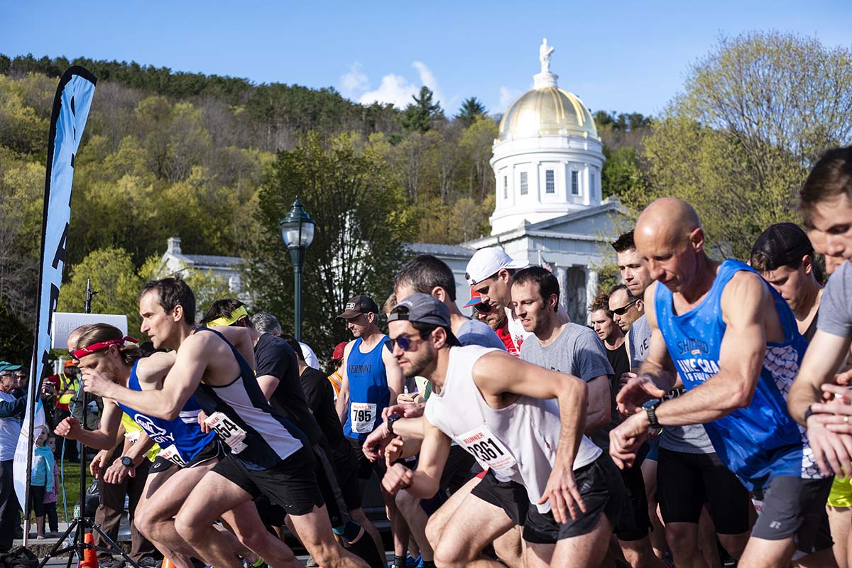 People wearing numbered race bib run past a sign that reads "start" with a view of Vermont's statehouse in the background